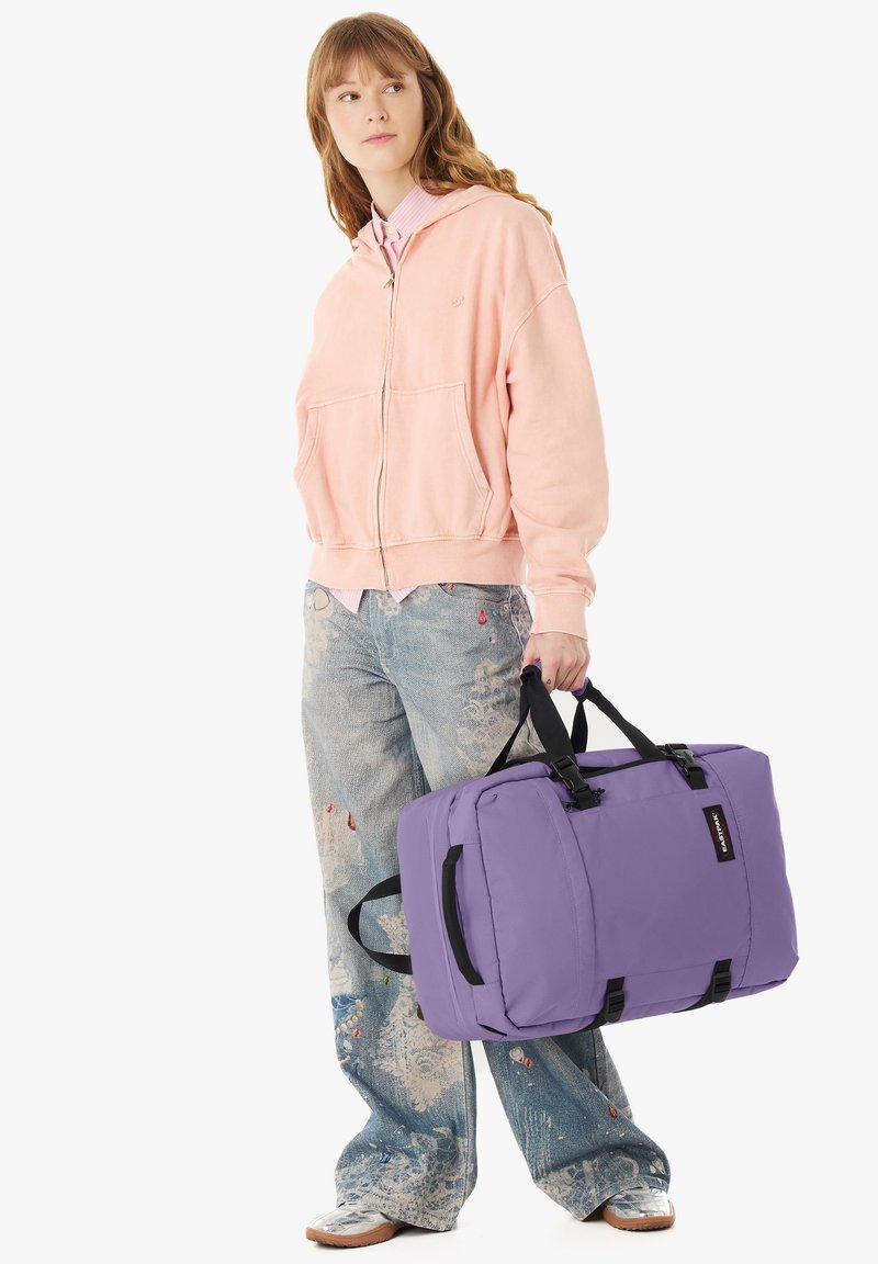 Jeune femme en sweat à capuche rose et jean à motifs tenant un grand sac de sport violet par ses poignées, debout devant un fond blanc.