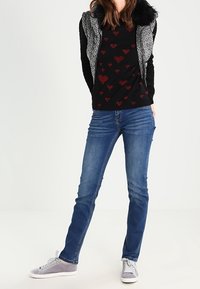 Zwarte trui met rode hartpatronen, grijze gewatteerde vest, blauwe skinny jeans en grijze sneakers met witte zolen. Over het algemeen een gladde textuur.