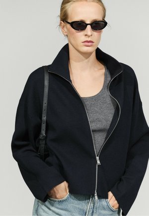 Donna che indossa una giacca nera con zip, top grigio a coste, jeans blu, occhiali da sole neri e porta una borsa nera a tracolla con le mani nelle tasche.
