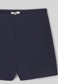 Shorts bleu marine en tissu lisse. Présentent une coupe droite, deux plis à l'avant et une fermeture éclair sur le côté. Design minimaliste.
