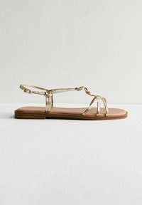 Gouden metallic platte sandalen met dunne bandjes en een geknoopte accent. Bruin gevoerd voetbed en een platte rubberen zool.