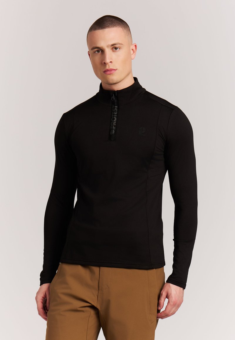 Giovane uomo con capelli corti che indossa una maglia nera con zip e maniche lunghe e pantaloni marroni, in piedi davanti a uno sfondo chiaro e uniforme.