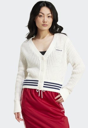Donna con lunghi capelli scuri che indossa un cardigan bianco lavorato a maglia con bottoni, con strisce navy, e una gonna rossa Adidas con strisce bianche.