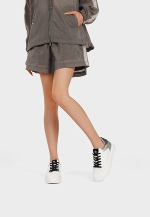 Persona che indossa una giacca sportiva grigia e pantaloncini con scarpe da ginnastica bianche con lacci neri e dettagli argentati sul tallone.