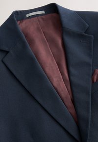 Blazer bleu marine texturé avec doublure intérieure à motif bordeaux et pochette assortie, présenté à plat sur un fond clair.