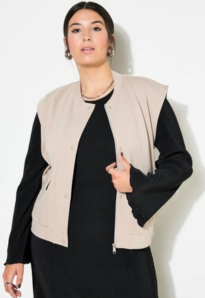 Beige bomber vest met een ronde hals, drukknoopsluiting en zijzakken, gelaagd over een zwart geribbelde jurk met uitlopende mouwen.