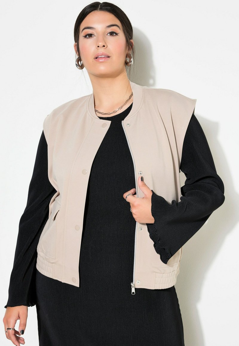 Beige Bomberweste mit rundem Ausschnitt, Druckknöpfen und Seitentaschen, über einem schwarzen gerippten Kleid mit ausgestellten Ärmeln getragen.