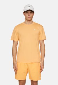 Orange atletisk t-shirt och shorts med vitt logotyp, korta ärmar och mjuk textur. Vit keps med svart logotyp fulländar outfiten.