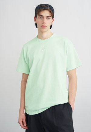 Junger Mann, trägt ein mintgrünes Nike-T-Shirt, schwarze Shorts und eine schwarze, rückwärts getragene Kappe, steht vor einem schlichten hellen Hintergrund.