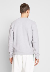 Uomo di spalle con un maglione grigio chiaro a maniche lunghe e pantaloni bianchi con una striscia verde di lato.