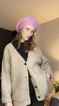 Bézs színű kötött kardigán laza szabással, nagy fekete gombokkal és két első zsebbel. Rózsaszín kötött barett sapkával párosítva.