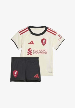 Dječja nogometna oprema Liverpool FC s kremastom majicom koja sadrži crvene logotipe i crnim hlačama s crvenim grbom Liverpoola i Adidasovim logotipom.