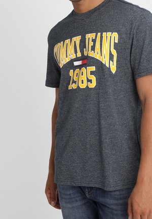 Camiseta de algodón gris con "TOMMY JEANS" y "1985" en letras amarillas, con un pequeño detalle en rojo y blanco. Mangas cortas, cuello redondo.