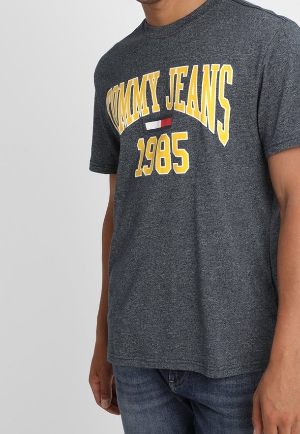 Camiseta de algodón gris con "TOMMY JEANS" y "1985" en letras amarillas, con un pequeño detalle en rojo y blanco. Mangas cortas, cuello redondo.