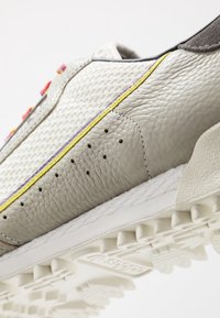 Grå läder- och mesh-sneakers med gul och lila sömnad, perforerade detaljer och en texturerad vit sula med greppande utsprång.