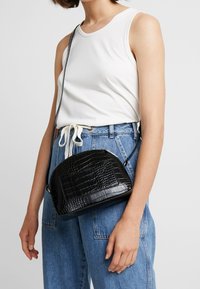 Borsa a tracolla nera con un motivo testurizzato in faux-coccodrillo, forma arrotondata, chiusura con zip e tracolla regolabile. Indossata con denim casual.