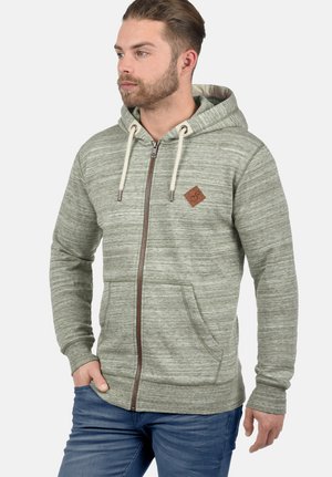 Lichtgroene ritshoodie van een gestructureerde stof, met crèmekleurige trektouwtjes, een bruine rits en een klein logopatch op de borst. Voorzakken zijn inbegrepen.