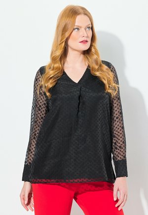 Schwarze Bluse mit Punkte-Muster, durchsichtigen langen Ärmeln, V-Ausschnitt und lockerer Passform, über einem einfarbigen roten Kleidungsstück getragen.