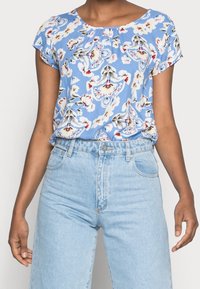 Bluse mit floralem Muster in Hellblau, kurzen Ärmeln und gerafftem Ausschnitt. Kombiniert mit hochgeschnittenen hellen Jeans.