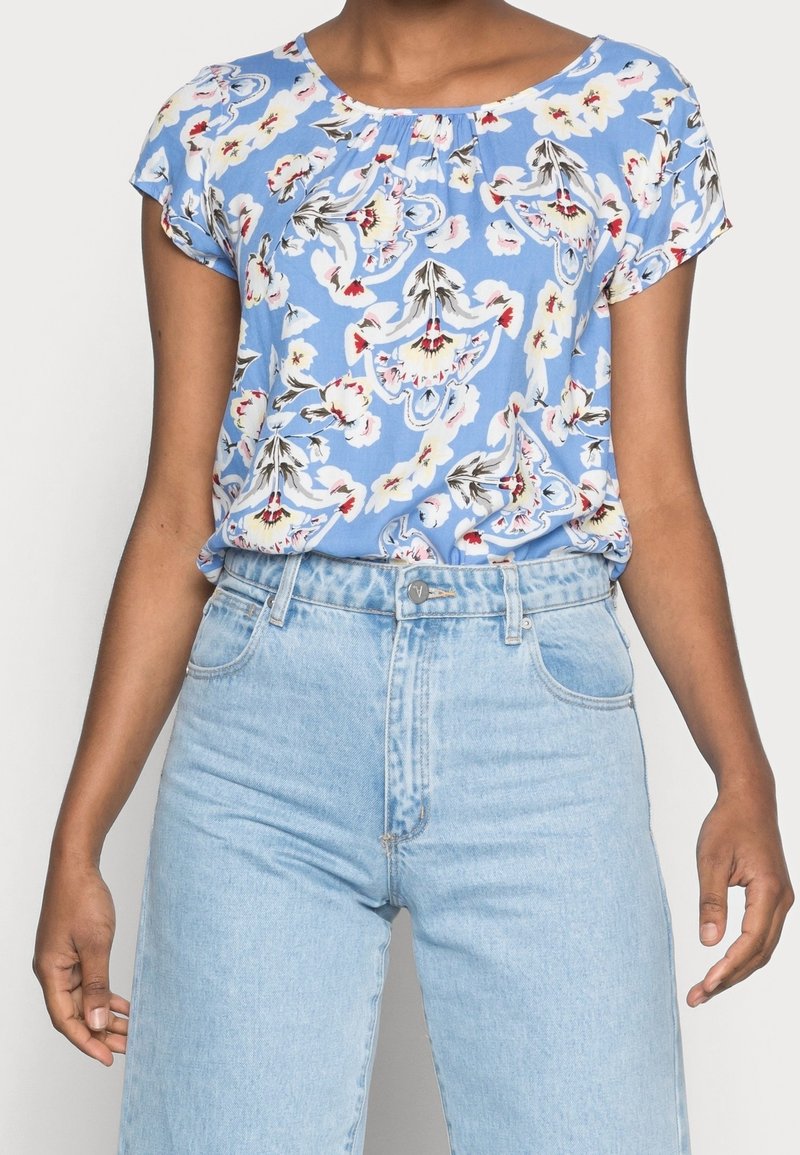 Bluse mit floralem Muster in Hellblau, kurzen Ärmeln und gerafftem Ausschnitt. Kombiniert mit hochgeschnittenen hellen Jeans.
