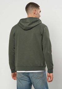 Mann in olivgrünem Hoodie und hellblauen Jeans steht mit dem Rücken zur Kamera vor einem schlichten hellen Hintergrund.