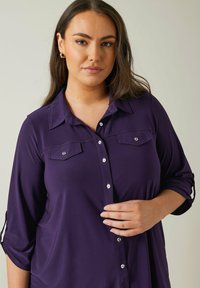Purple shirt with button-down front, two chest pockets, and rolled 3/4 sleeves. Made from a smooth fabric with subtle sheen.