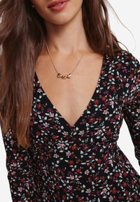 Blommönstrad klänning i svart tyg med V-ringning, prydd med små röda och vita blommor; kompletteras med ett guldhalsband med länkade hjärtan.