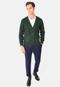Cardigan verde lavorato a maglia con scollo a V, texture a coste, due tasche frontali e bottoni neri, abbinato a pantaloni blu navy e scarpe nere.