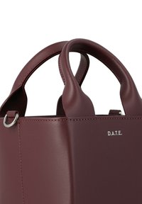 Donker bordeauxleren handtas met ronde handvatten en een zilveren ring, met kleine witte merkinitialen "D.A.T.E." nabij de bovenrand.