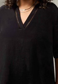 Primo piano di una persona che indossa una maglietta nera a V con rifiniture in rete e collane d'argento sovrapposte.