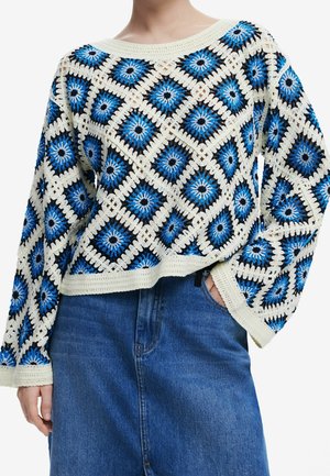 Vrouw draagt een gehaakte trui met blauwe, zwarte en witte ruitpatronen, gecombineerd met een high-waisted blauwe spijkerbroek.