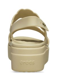 Sandali Crocs color crema con suola spessa e testurizzata, con cinturini incrociati e un design scallopato sul retro. Cinturino regolabile con dettaglio in metallo.
