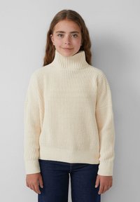 Crème-kleurige gebreide trui met een hoge kraag, geribbelde textuur, een relaxte pasvorm en wijde mouwen; gecombineerd met donkere jeans.