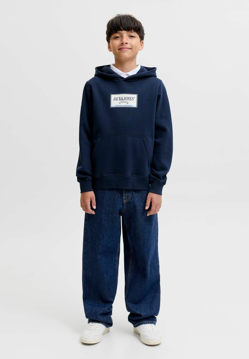 Garçon portant un sweat à capuche bleu marine avec le logo Jack & Jones, un jean ample foncé et des baskets blanches, debout devant un fond clair uni.