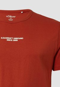 Rustfarbenes Baumwoll-T-Shirt mit Rundhalsausschnitt, kurzen Ärmeln und weißem Text "S.OLIVER® UNBOUND SINCE 1969" auf der Vorderseite.