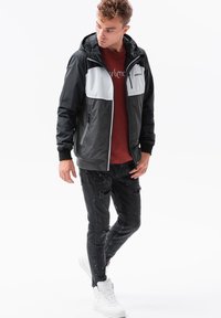 Giacca leggera nera e grigia con cappuccio, tasche con zip e polsini a costine, abbinata a jeans neri strappati e sneaker bianche.