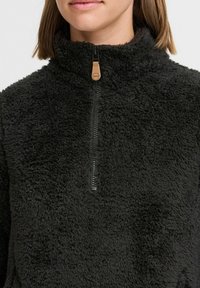 Černý fleecový pulovr s vysokým límcem a uzávěrem na půl zip. Má měkký, texturovaný povrch a hnědý kožený táhlo.