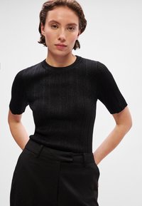 Juodas trumpų rankovių megztinis su apvaliu iškirpimu. Turi tekstūrinį paviršių ir aptemptą siluetą. Dera su pritaikytos juodos spalvos kelnėmis.