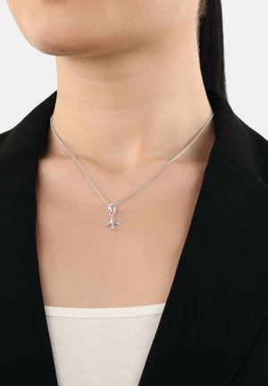 Silberner Flugzeuganhänger-Halskette auf einer feinen Kette, die auf einem neutralen Oberteil ruht, mit einem schwarzen Blazer im Hintergrund.