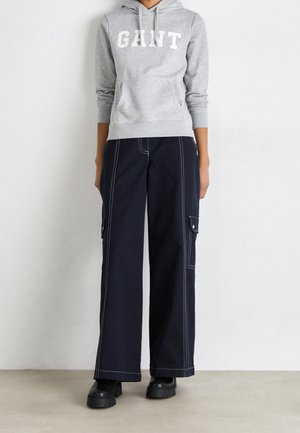 Personne portant un sweat à capuche gris GANT, un pantalon cargo large noir avec des coutures blanches et des bottes à plateforme noires, debout contre un mur uni.