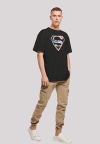 Black cotton T-shirt with a colorful Superman logo, paired with beige cargo pants. Model wears white sneakers. Casual fit and design.