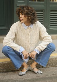 Cardigan en tricot beige clair avec un motif en relief et des boutons dorés, porté sur une chemise col bleu clair. Jean flare bleu et ballerines vichy noir et blanc.
