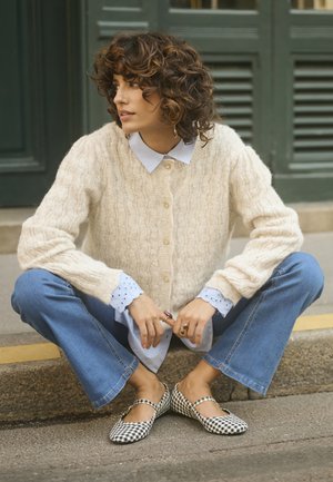 Light beige cable-knit cardigan with gold buttons, worn over a light blue collared shirt. Blue flared jeans and black-and-white gingham flats.