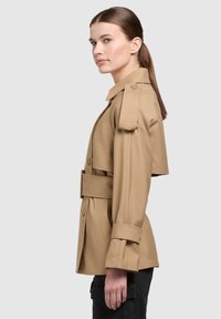 Beżowy trench coat z szerokim kołnierzem, z paskiem w talii i naramiennikami. Gładki materiał, dopasowany krój oraz guziki na mankietach.