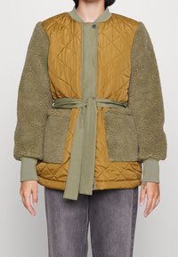 Kvinna klädd i en brun vadderad jacka med olivgröna fleecesleeves och matchande bälte knutet i midjan, i kombination med grå byxor.