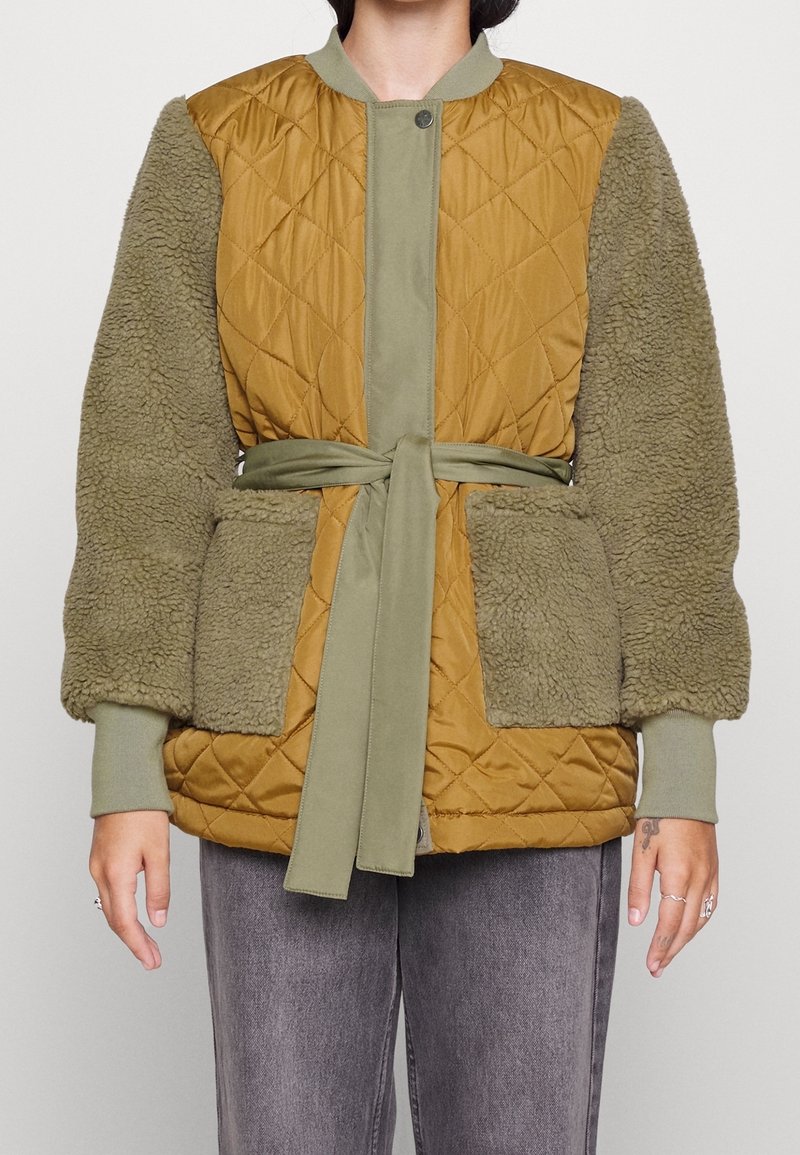 Kvinna klädd i en brun vadderad jacka med olivgröna fleecesleeves och matchande bälte knutet i midjan, i kombination med grå byxor.