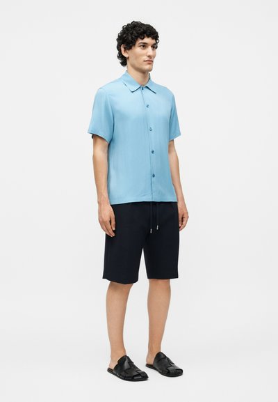 Νέος άνδρας στέκεται φορώντας ένα ανοιχτό μπλε πουκάμισο με κοντά μανίκια και κουμπιά, μαύρα σορτσάκια με κορδόνι και μαύρα σανδάλια με ανοιχτά δάχτυλα.