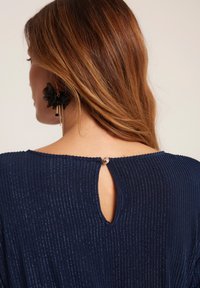 Granatowa sukienka z pionową teksturą ryżu, z dekoltem w kształcie klucza z tyłu, zabezpieczonym okrągłym guzikiem, noszona z czarnym kwiatowym kolczykiem.