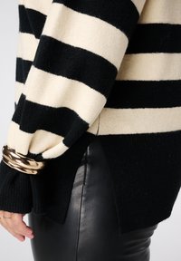 Pull rayé noir et crème à coupe ample, poignets côtelés et fentes latérales, associé à un pantalon en cuir noir ajusté. Détails en métal doré.