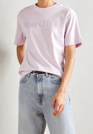 Osoba nosząca jasnofioletowy t-shirt "Marc O'Polo" włożony do jasnoniebieskich, wysokich spodni dżinsowych z czarnym paskiem.